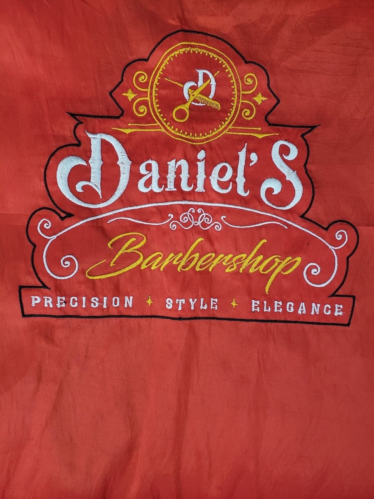 The Daniel’s Barbershop logo embroidered in gold and white on a red barber vest