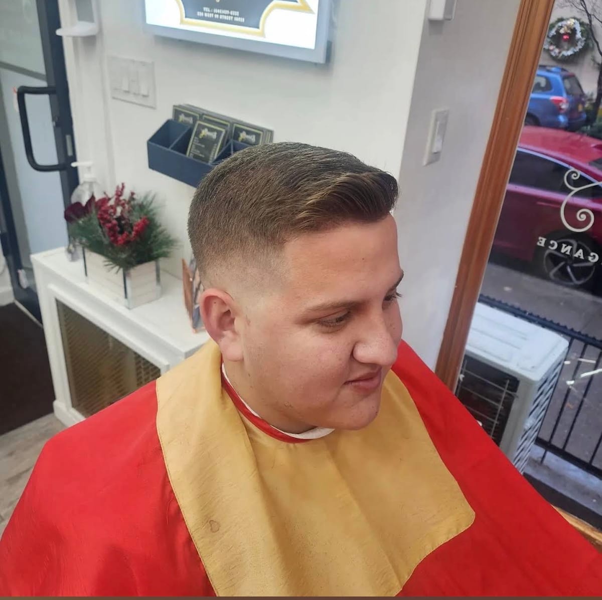 Side view of a short fade with a swept-back top on a client in a red and gold cape, the shop window and storefront behind him